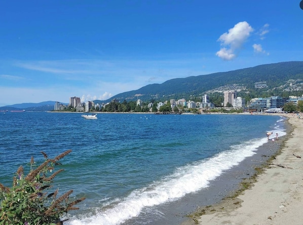 A coastal cityscape with a beach, buildings, and a boat on the water. - West Van Luxury Retreat 2BR 2BA Oceanview (West Vancouver)