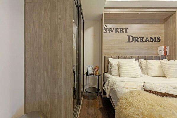 Ein Schlafzimmer mit einem Bett, einem Nachttisch mit Büchern, einem flauschigen Teppich und einer Wand mit der Aufschrift 'Sweet Dreams'.