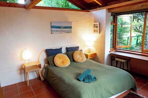 Ein Schlafzimmer mit einem Bett, zwei Nachttischlampen, einem Holznachttisch, einem Fenster mit Blick ins Grüne und einem Wandbild.