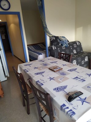 Uma sala de jantar com uma mesa coberta por toalha azul e branca, cadeiras de madeira, um relógio na parede e uma cama com almofadas estampadas.