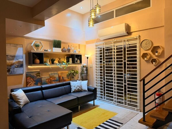 A living room with a black sofa, a yellow rug, a staircase, and a white-framed window with blinds.