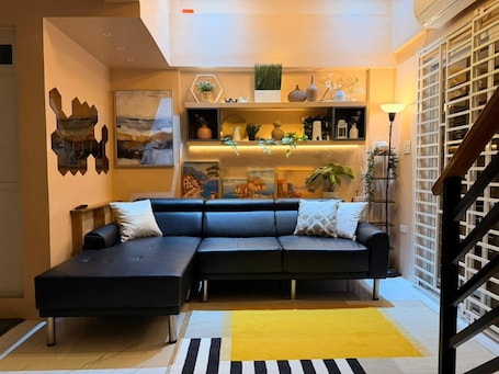 A modern living room with a sectional sofa, a yellow rug, and decorative shelves.. Welcoming 1-bedroom apartment with AC in charming Lucban