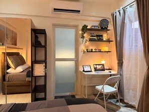 A bedroom with a bed, a desk, a chair, a shelf with books, a clock, and a window with curtains.