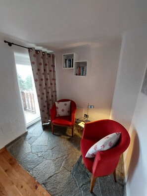 Ein Zimmer mit Steinboden, zwei roten Stühlen, einem kleinen Tisch, einer Stehlampe und einer Schiebetür mit floralem Vorhang.