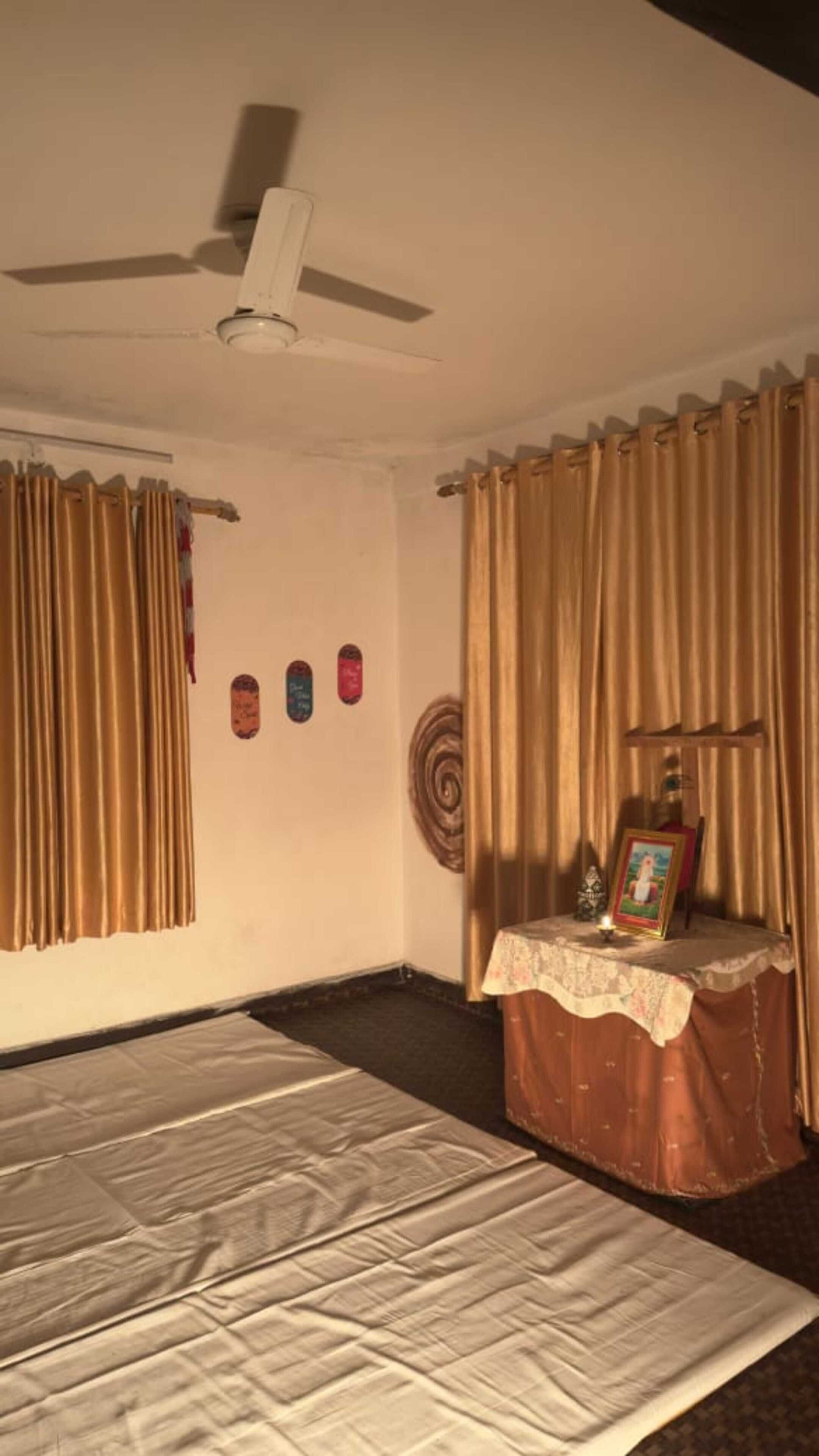 A room with a bed, a ceiling fan, curtains, a small table with a picture, and wall decorations.