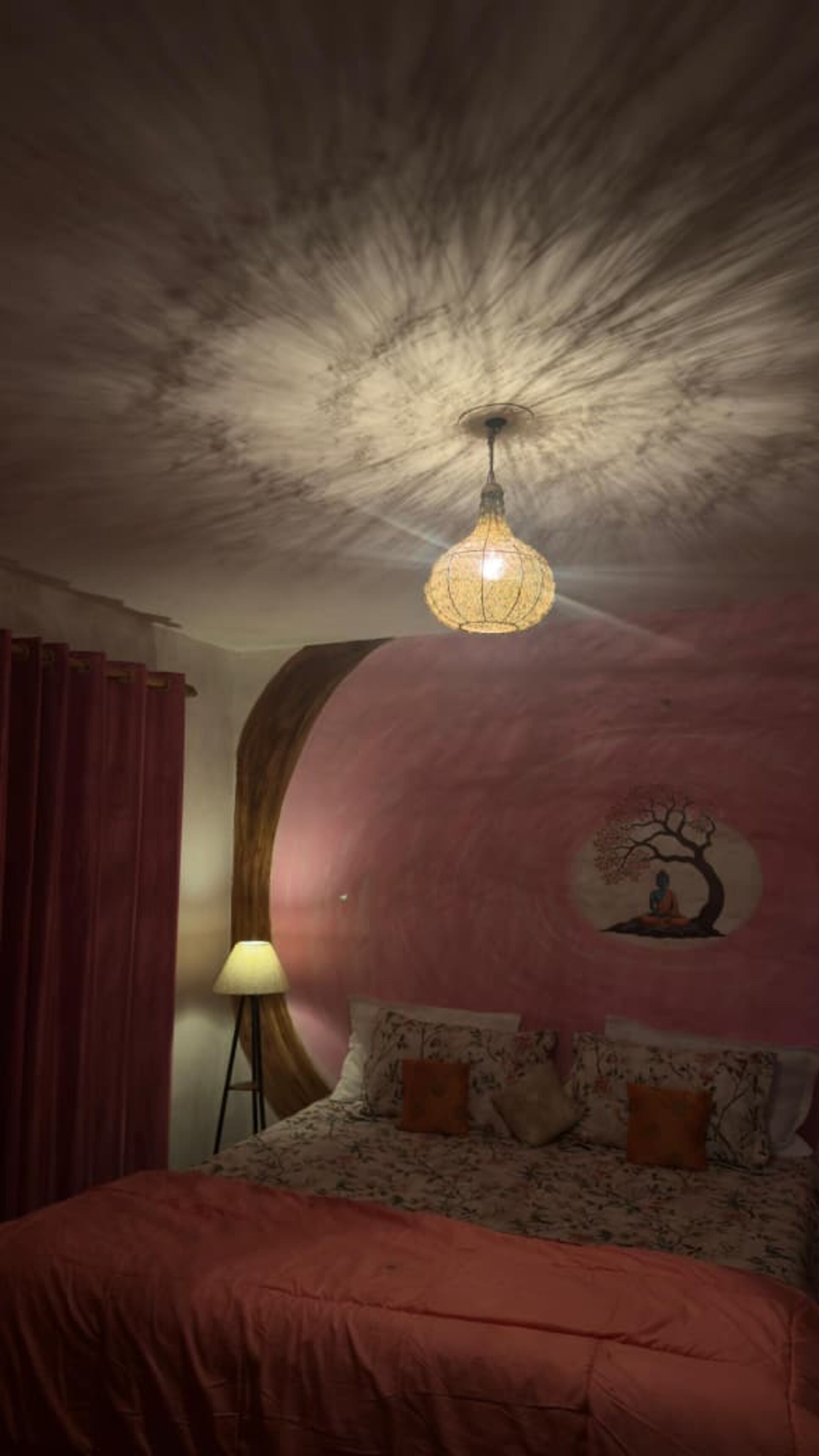 Un dormitorio con cama, lámpara, luz de techo y un muro con un árbol y una figura.