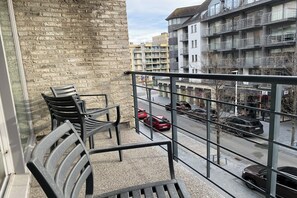 En balkon med stole, udsigt over en gade med biler, og en bygning med balkoner.