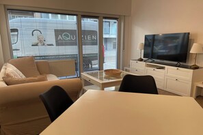 A modern living room with a sofa, a dining table, a TV, and a window with blinds. - Coastal Retreat in Nieuwpoort-Bad (NIEUWPOORT)