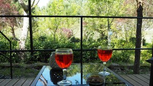 Una mesa con dos copas de vino y una botella, sobre un fondo de follaje y árboles.