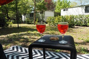 Zwei Weingläser auf einem Tisch mit gemustertem Tischläufer, umgeben von Grünpflanzen und einem weißen Gebäude im Hintergrund.