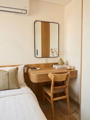 Habitación de hotel pequeña con cama, un escritorio de madera, una silla, un espejo, una mesita de noche y una planta pequeña.