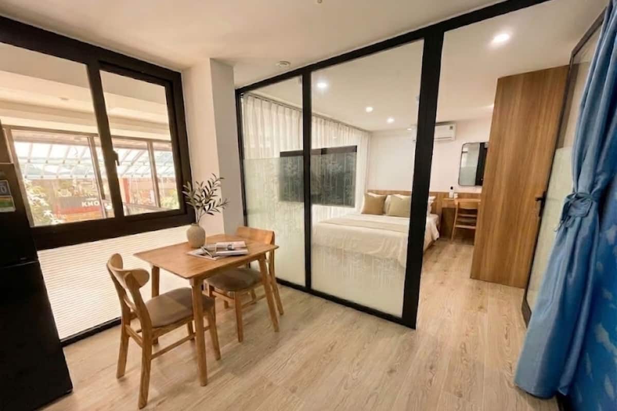 A modern room with a wooden floor, a dining table with chairs, a bed with a white duvet, and a sliding glass door leading to another room.