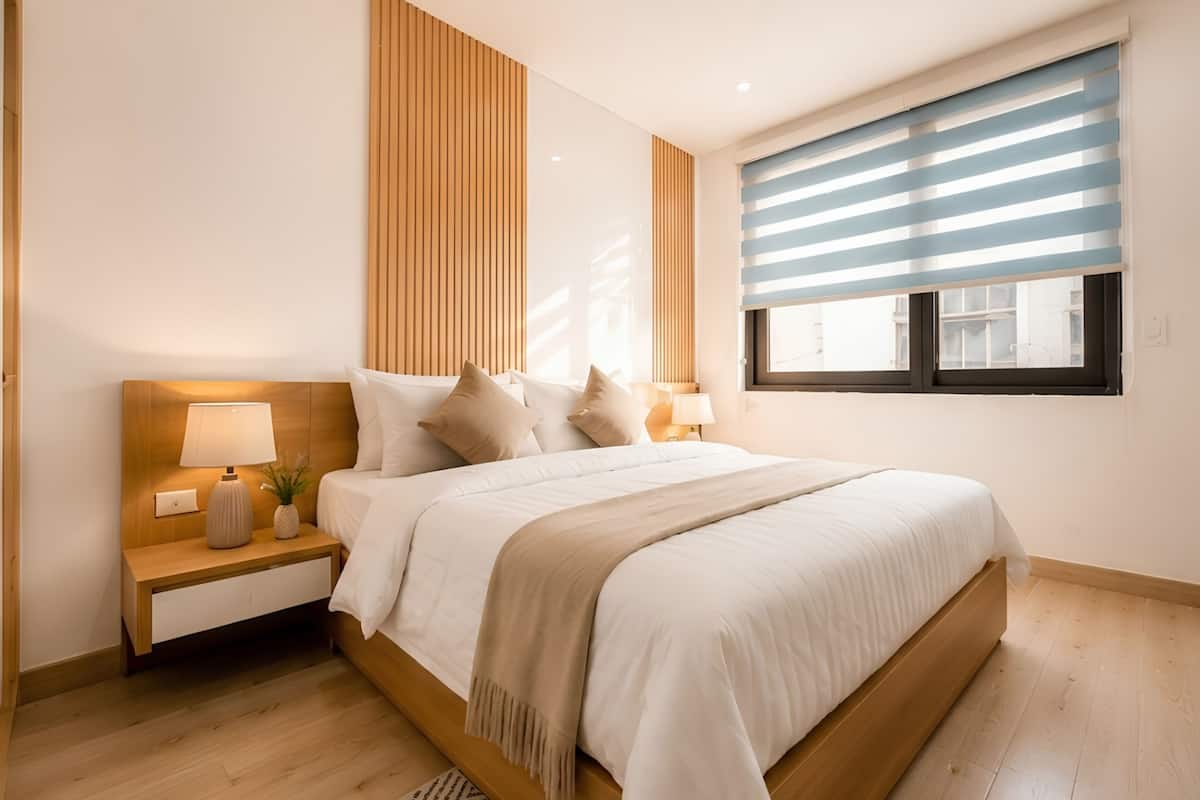 A modern bedroom with a wooden headboard, a large bed, bedside tables with lamps, and a window with horizontal blinds.