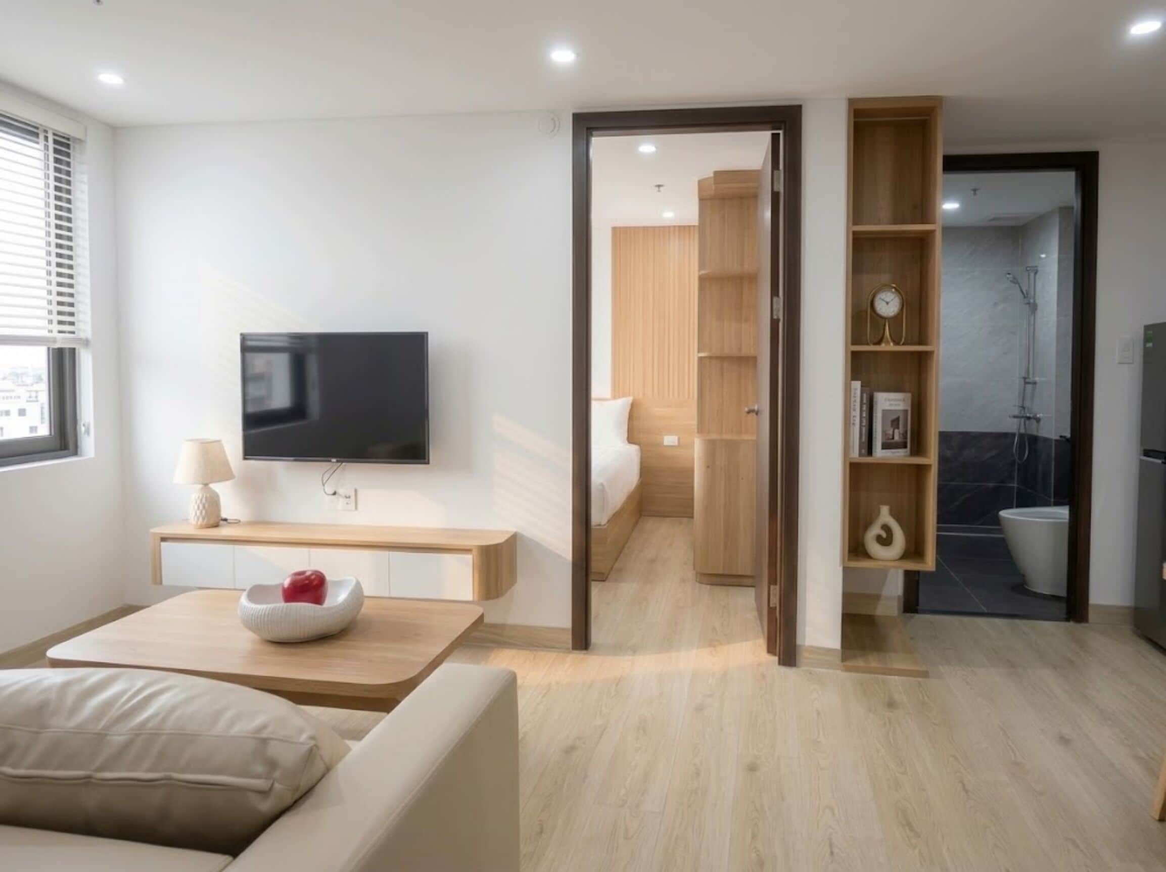 Una habitación de hotel moderna con una televisión de pantalla plana, una mesita de noche con lámpara, un frutero con una manzana, un sofá y suelo de madera.