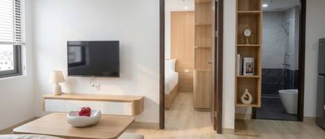 Una habitación de hotel moderna con una televisión de pantalla plana, una mesita de noche con lámpara, un frutero con una manzana, un sofá y suelo de madera.