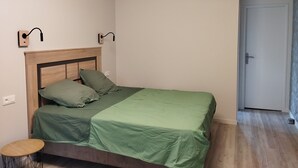 A single bed with a wooden headboard, green bedding, two wall-mounted light fixtures, a small round table, and a door to the right. - La Canissa Carrada family canopy room (Chaspuzac)