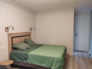 Una cama individual con cabecera de madera, ropa de cama verde, dos luces fijadas a la pared, una mesita redonda y una puerta a la derecha.