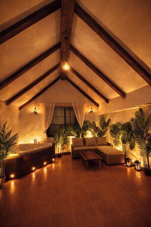 A spacious room with a sloped ceiling, wooden beams, a large window with curtains, a bathtub, a sofa, a coffee table, and several potted plants. - 500m2 Jacuzzi House 6BR West Lake/Billiard/BBQ (Hà Nội)