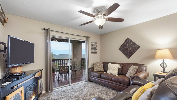 A living room with a fireplace, a ceiling fan, a television, a couch, and a view through a glass door to an outdoor patio. - View of Table Rock Lake - Minutes from SDC - PERFECT! (Branson)
