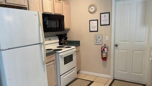 Una cucina con frigorifero, fornello e microonde bianchi, mobili in legno, una sveglia, certificati incorniciati e un estintore.