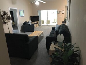 Ein Wohnzimmer mit einer Couch, einem Couchtisch und einem Fernseher. Eine Pflanze und ein Deckenventilator sind ebenfalls vorhanden.