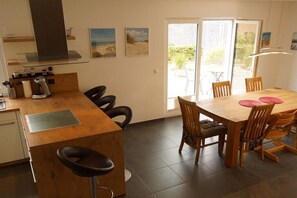 Une cuisine moderne avec un îlot central en bois, des tabourets de bar et une table à manger avec des chaises. Il y a deux fenêtres équipées de stores et des œuvres d’art accrochées aux murs.