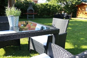 A glass-top outdoor table with wicker chairs, surrounded by a grassy lawn and a barbecue grill. - Carefree House Mini-House (Travemünde)