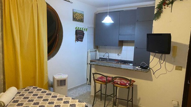 Een compacte kamer met een bed, een kleine kitchenette, twee barkrukken, een televisie en een plafondventilator.