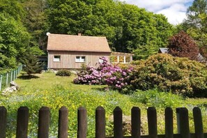 A wooden house with a brick roof, surrounded by lush greenery and flowering bushes. - Holiday home with 2 bedrooms (Kurort Oybin OT Lückendorf)