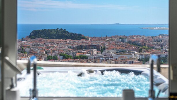 Une salle de bain avec une fenêtre donnant sur un paysage urbain côtier, comprenant une colline et la mer.
