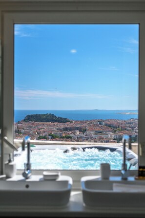 Une salle de bain avec une fenêtre donnant sur un paysage urbain côtier, comprenant une colline et la mer.