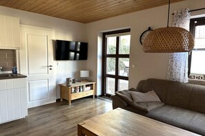 Ein Wohnzimmer mit einer Couch, einem Holztisch, einem an der Wand montierten Fernseher und einem Fenster mit Vorhängen.