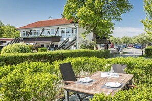 Ein Holztisch im Freien mit Stühlen, ein weißes Gebäude mit rotem Dach und ein gepflegter Hecken im Vordergrund.