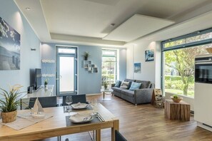 Ein modernes Wohnzimmer mit Essbereich, großen Fenstern und einer durchgehenden Tür erkennbaren Küche.
