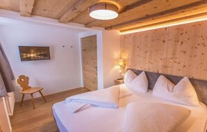 Ein Schlafzimmer mit Holzboden, einem Bett mit weißer Bettwäsche, einem Sessel, einem an der Wand montierten Fernseher und einem Nachttisch mit Lampe.