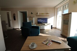 Ein modernes Wohnzimmer mit Fernseher, einer Couch, einem Couchtisch mit Fernbedienung und einer Tasse sowie einer Tür zu einem weiteren Raum.