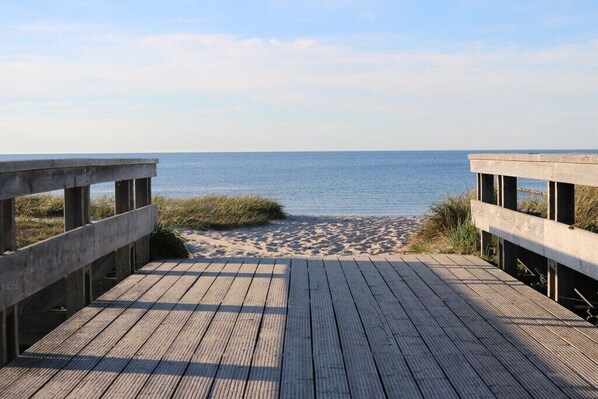 Ein hölzerner Steg führt zu einem Sandstrand, im Hintergrund das Meer.