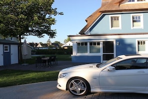 Ein wei ßes Auto vor einem blau gestrichenen Haus mit Strohdach, einer Terrasse mit Stühlen und einem Rasen.