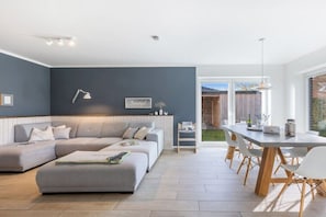 Ein modernes Wohnzimmer mit einem grauen Ecksofa, einem Esstisch mit Stühlen und einer kleinen Küchenzeile.
