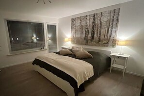 A bedroom with a bed, two bedside lamps, a window with blinds, and a large forest-themed painting on the wall. - Beach house connected to the sea (Kellenhusen)