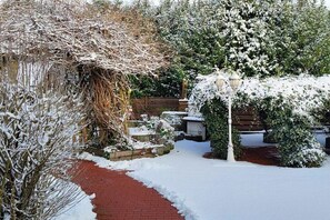 A snow-covered garden with a brick pathway, a wooden fence, and a stone wall. - Holiday apartment with 2 bedrooms (21706 Drochtersen, Deutschland)