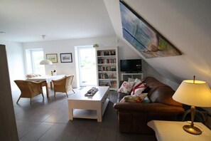Ein Wohnzimmer mit einer Couch, einem Couchtisch und einem Fernseher. Ein Bücherregal und ein Essbereich mit Stühlen und einem Tisch sind ebenfalls vorhanden.