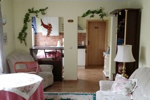 Ein Wohnzimmer mit einer Couch, einem Tisch und einer Lampe. Es gibt eine Küchenzeile mit Schränken und eine Tür zu einem anderen Raum.