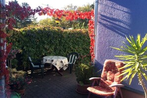 En udendørs terrasse med et bord og stole, omgivet af grønne planter og en mur.