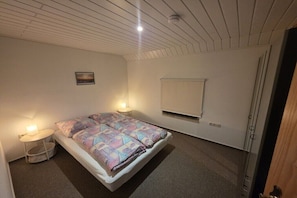 Ein Einzelbett mit gemustertem Überwurf, ein Nachttisch mit Lampe, eine kleine Wandablage und ein Bild an der Wand.