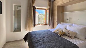 Ein Schlafzimmer mit Bett, einem fensterartigen Durchblick mit Vorhängen, einem Spiegel und Blick auf Gebäude.