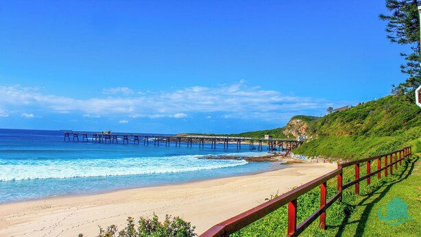 A beach with a pier extending into the ocean, a grassy hill, and a red railing along the shore. - Wagtail Whispers - Spacious Coastal Escape (Catherine Hill Bay)