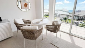 A balcony with wicker chairs, a round table, and a large window offering a view of buildings and greenery. - Wagtail Whispers - Spacious Coastal Escape (Catherine Hill Bay)