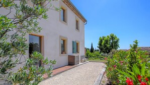 A two-story house with a gravel driveway, wooden deck, and a garden with various plants and flowers. - Bastide Néo Provencale (Grignan)