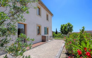 A two-story house with a gravel driveway, wooden deck, and a garden with various plants and flowers. - Bastide Néo Provencale (Grignan)